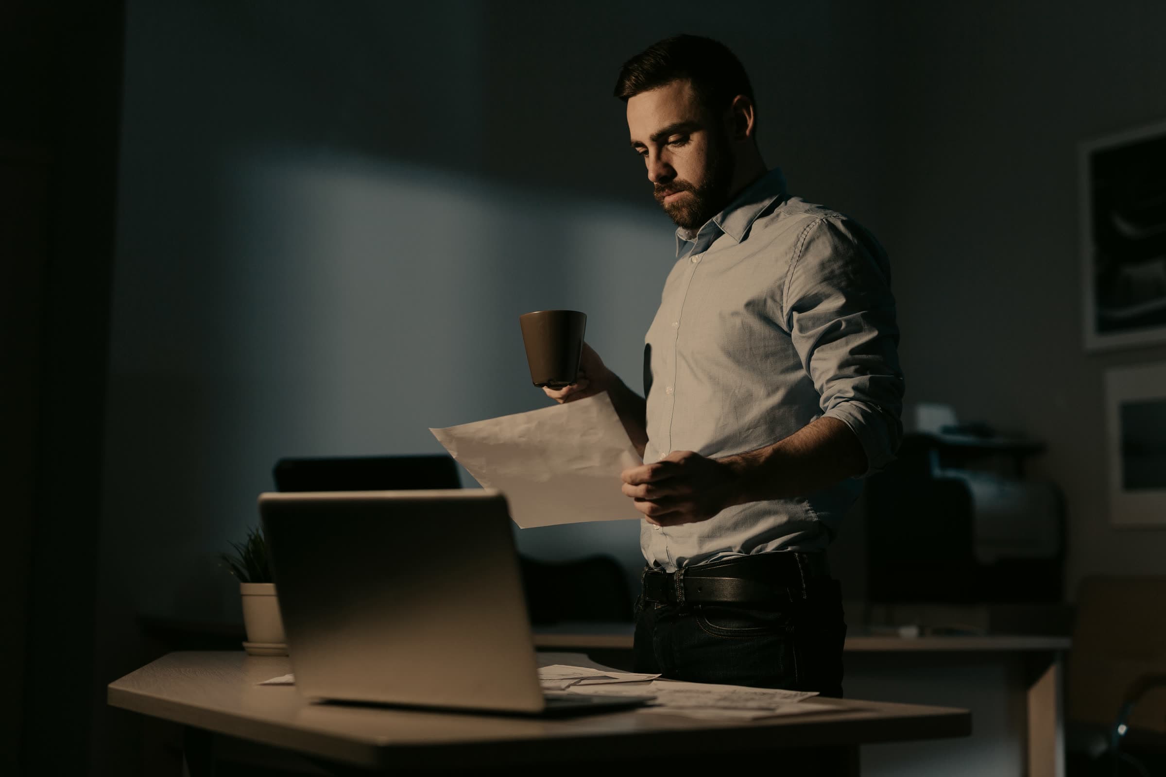 Man with a coffee reading printed documents at a desk in a dark office at night