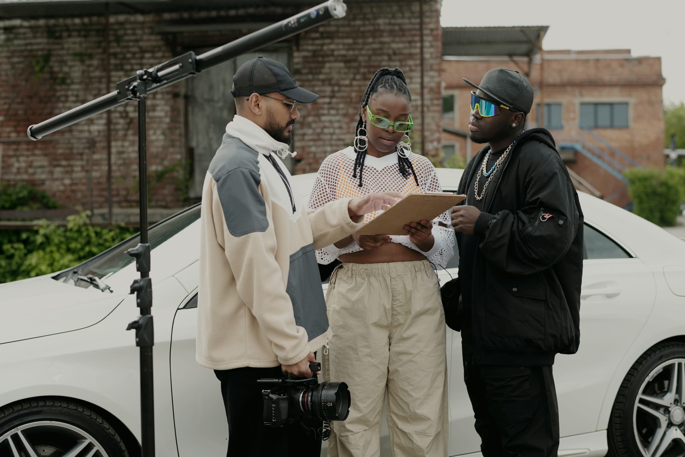 Crew member walking talent and a colleague through a clipboard beside a car on a location shoot, boom stand overhead
